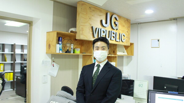 제이지비퍼블릭 로고 앞에서 김재관 대표가 웃고있는 모습